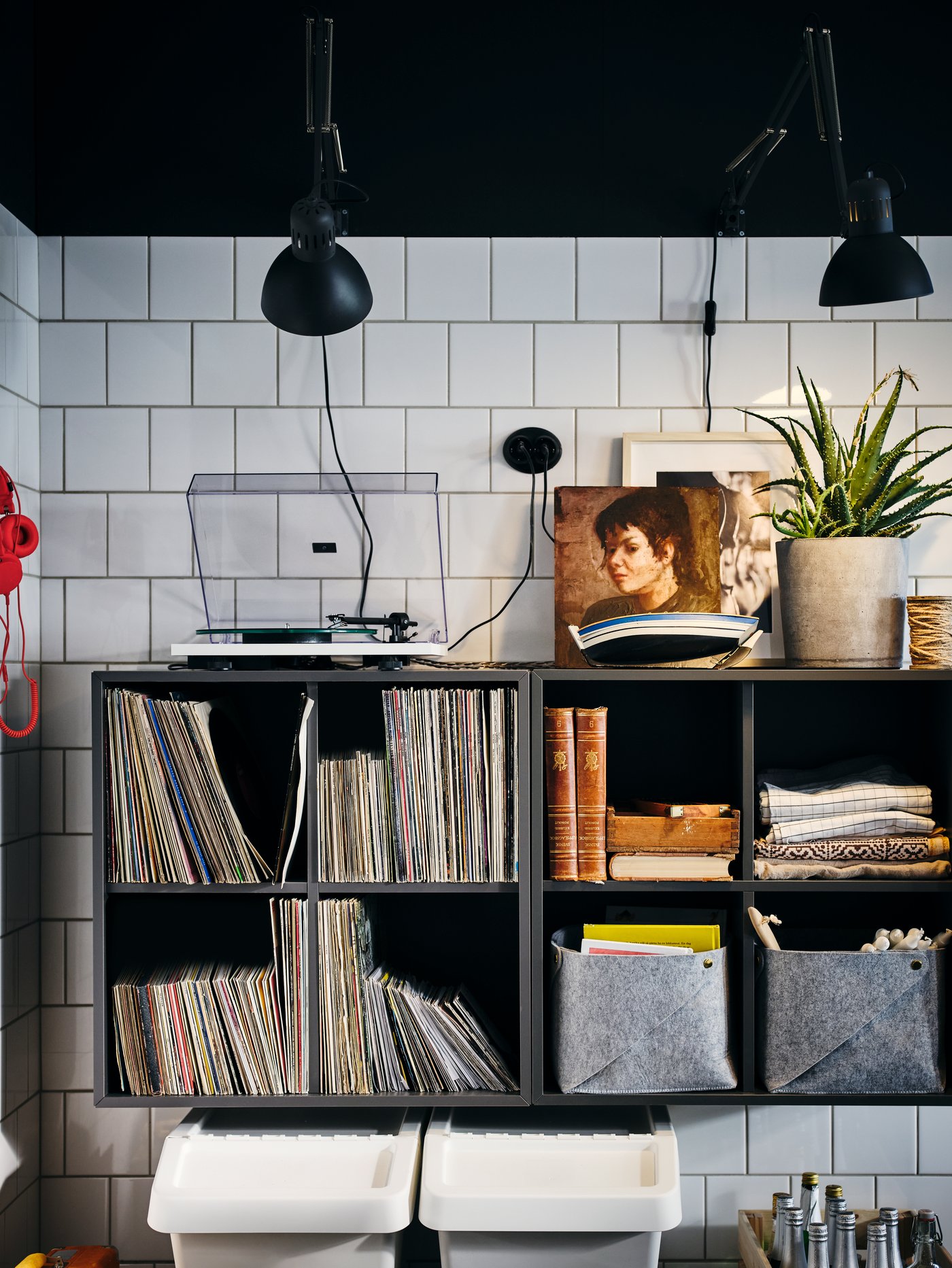 A corner of a white-tiled kitchen with lamps and side-by-side, wall-mounted, dark-grey EKET four-compartment cabinets.