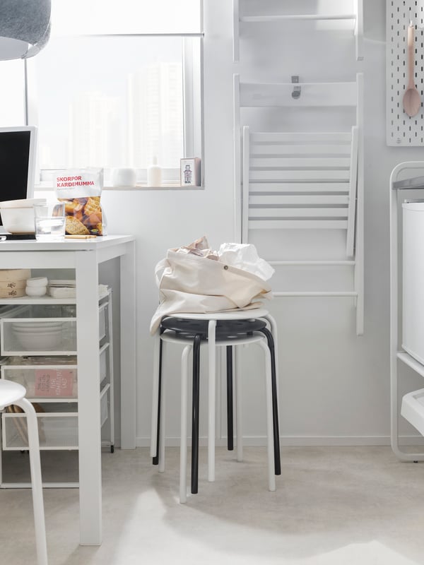 A kitchen corner with a white table and white folding chairs hanging on the wall, and stools stacked in front.