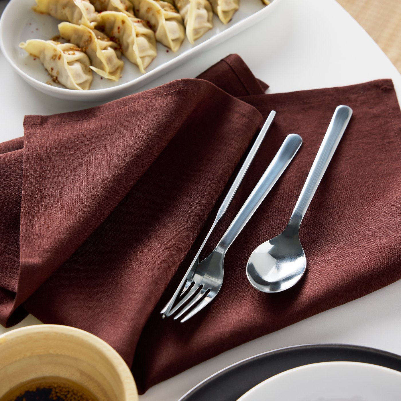 Una forchetta, un coltello e un cucchiaio della serie di posate IKEA 365+ sono appoggiati su un tovagliolo STOCKHOLM 2025 rosso scuro.