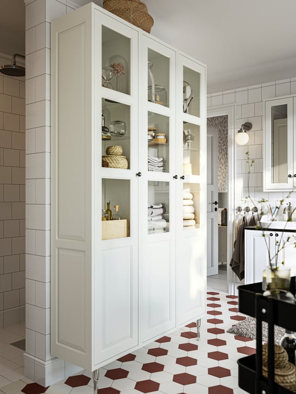 A bathroom featuring a white TÄNNFORSEN high cabinet with glass doors filled with towels, boxes and bottles.
