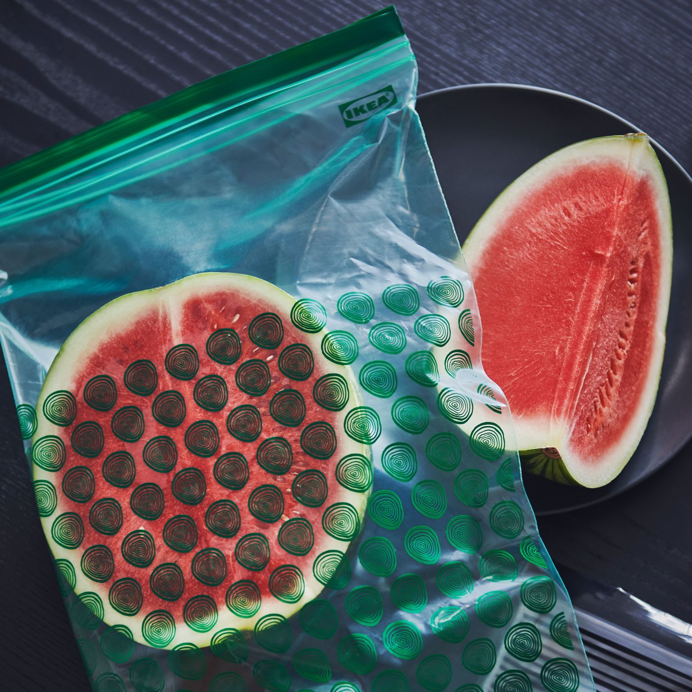 Half a watermelon in an ISTAD resealable bag with a green pattern is placed next to a plate with another slice of watermelon.