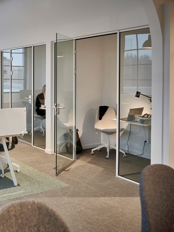 Små privata kontor med glasväggar i öppet kontorslandskap innehållande litet väggfast skrivbord och skrivbordsstol. 