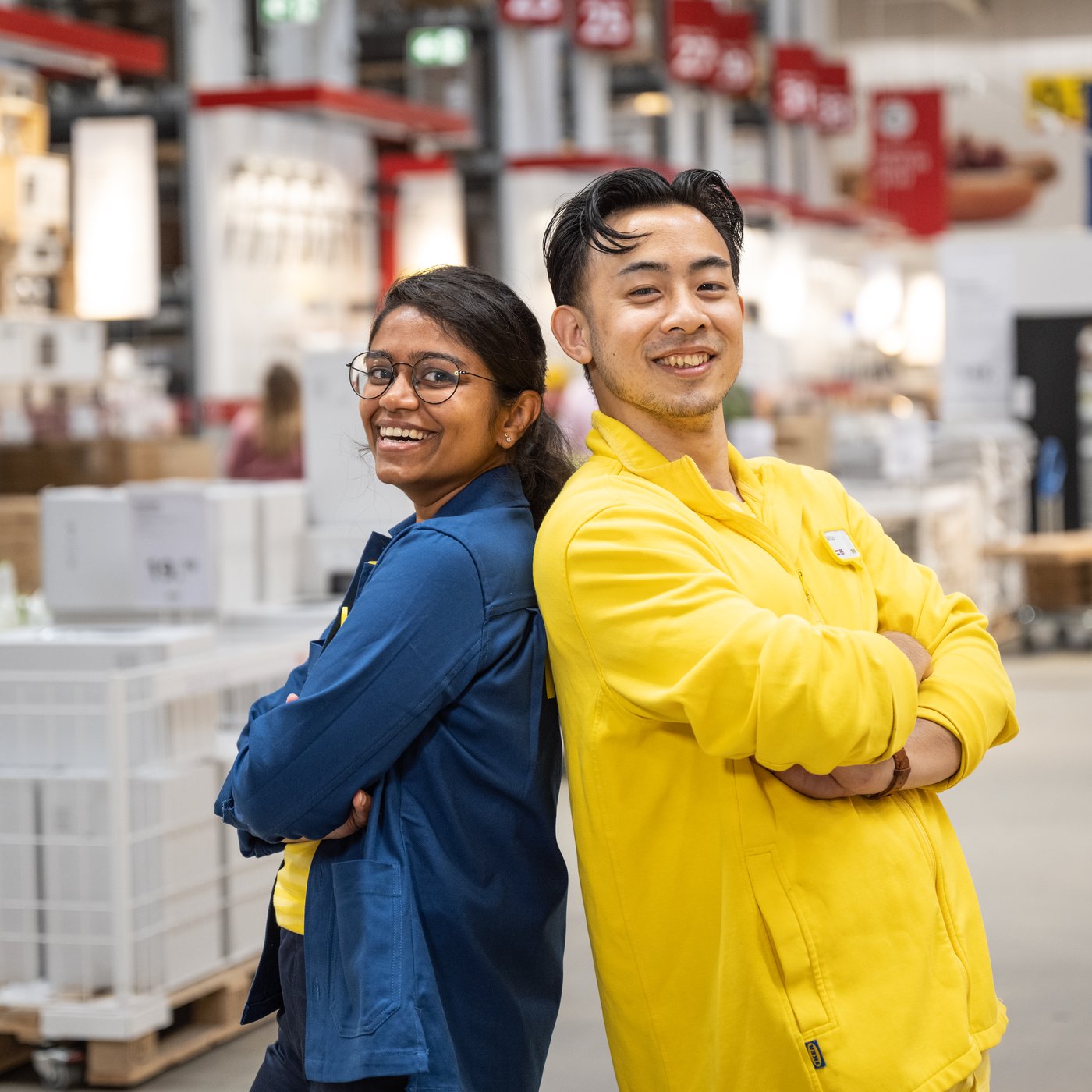 Twee IKEA medewerkers staan met hun rug tegen elkaar in een magazijn. Ze dragen een IKEA werkoutfit in blauw en geel. Op de achtergrond staan rekken en pallets.