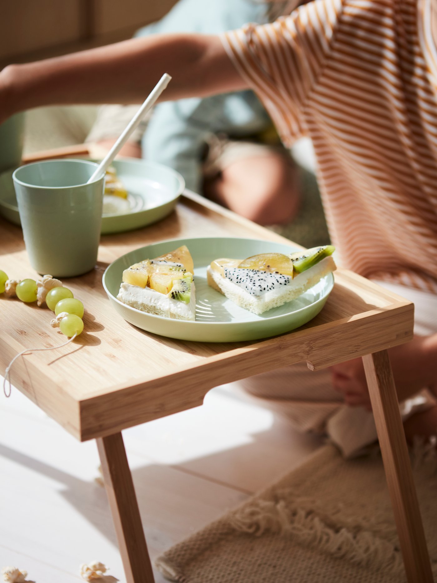 A close-up of a bambo bed tray standing on the floor holding a fruity snack on green KALAS dinnerware with a child behind.