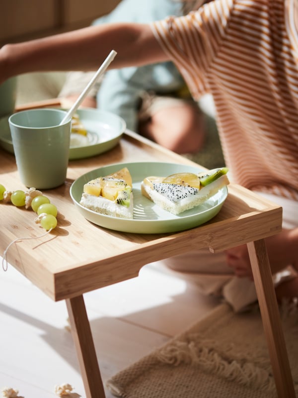 A close-up of a bambo bed tray standing on the floor holding a fruity snack on green KALAS dinnerware with a child behind.