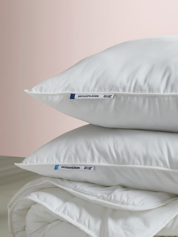 Twee SKOGSFRÄKEN kussens, een hoog en een laag, liggen gestapeld op een dekbed tegen een zachte pastelkleurige achtergrond.
