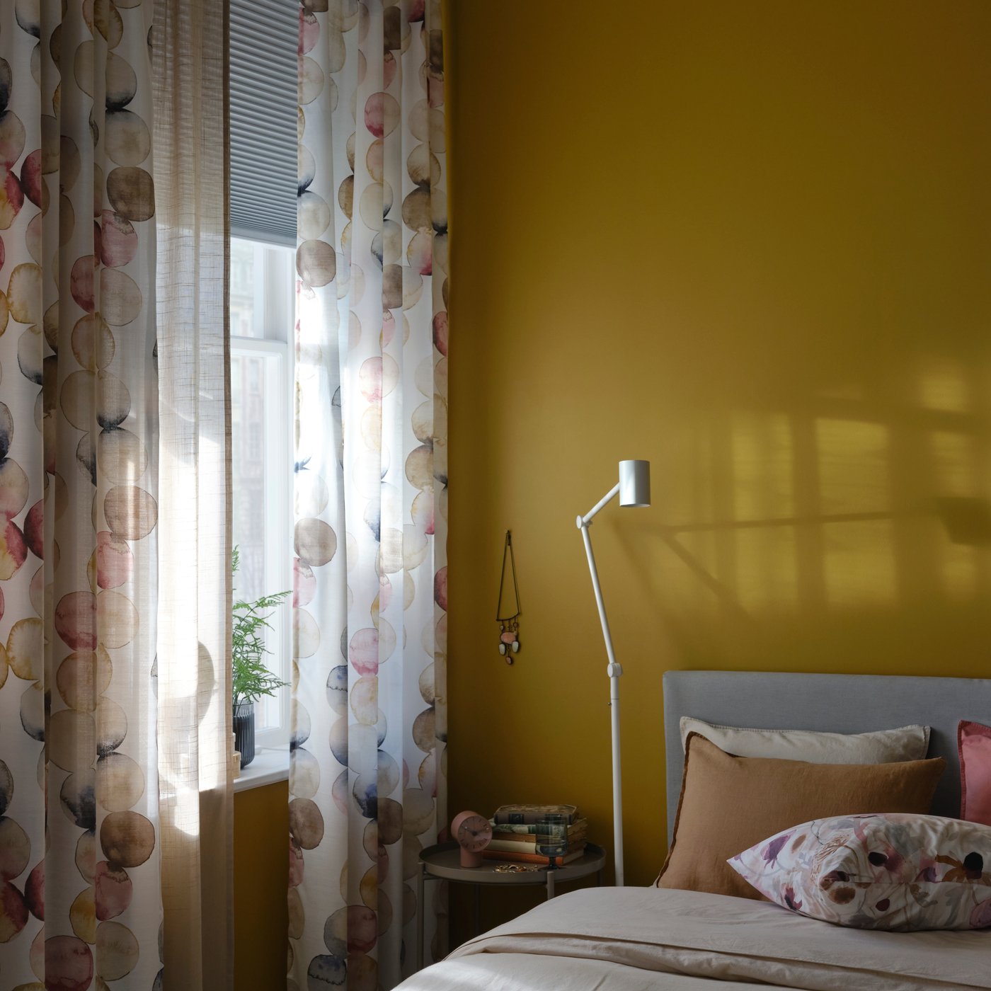 Un lit et un lampadaire/liseuse blanc près d’une fenêtre avec des rideaux multicolores SÄCKMAL et un store alvéolaire occultant.