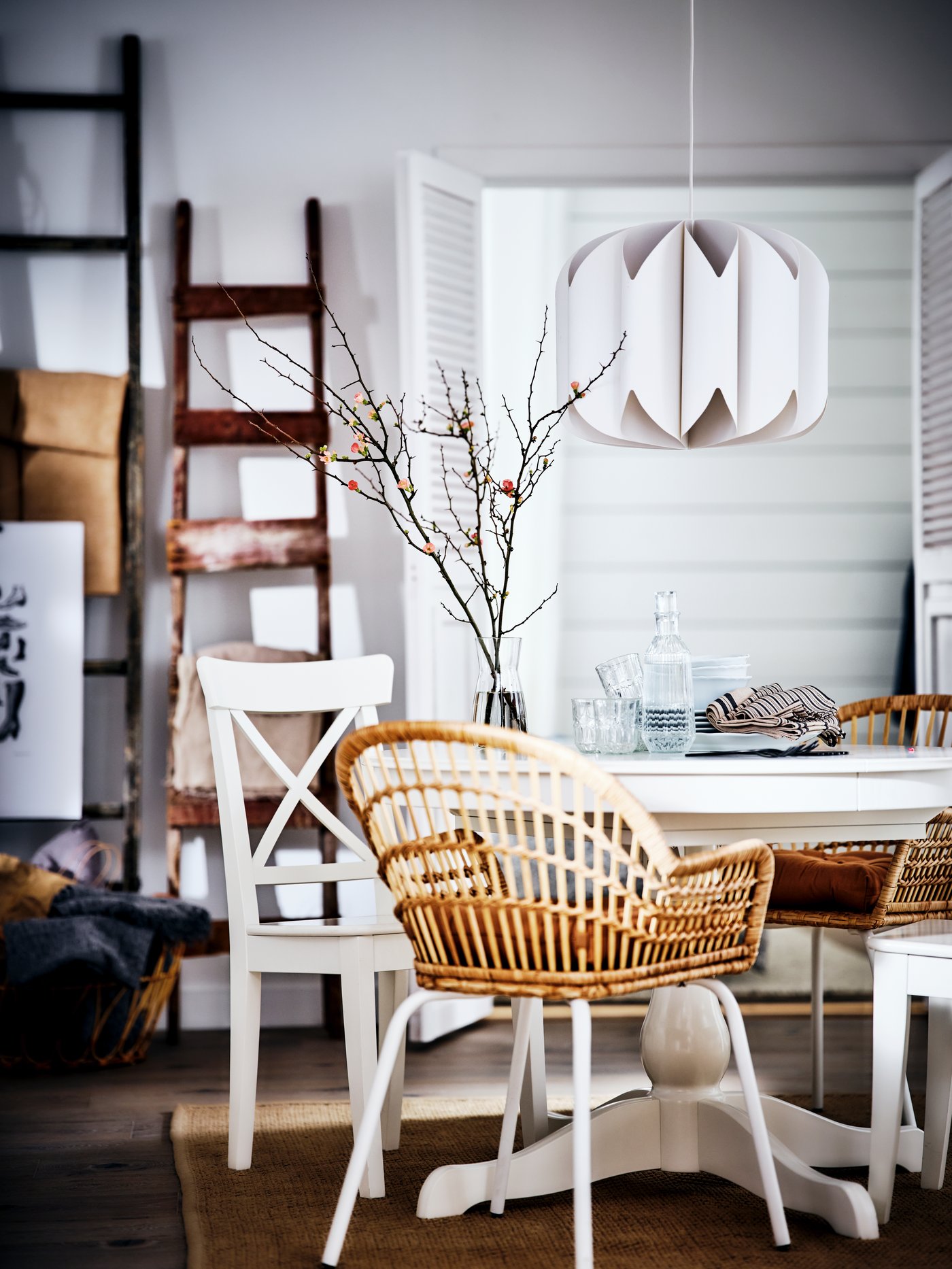 A white table with a mix of rattan and wooden chairs around it, and with crockery and glasses on top in a white dining room.