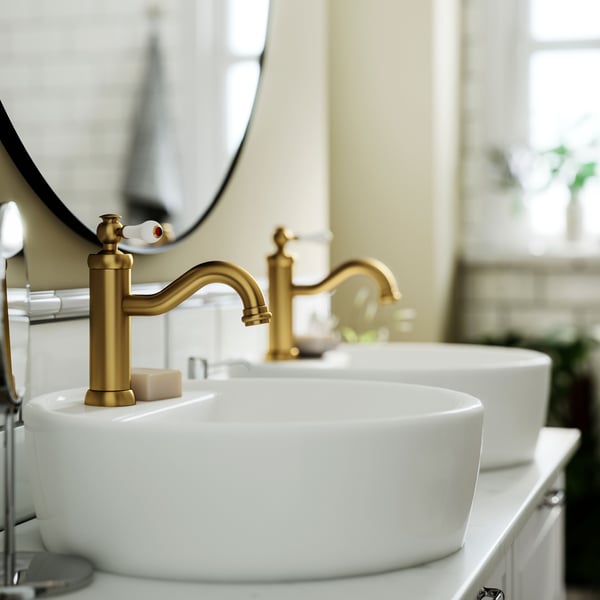 Dans une pièce claire, deux mitigeurs pour lavabo HAMNSKÄR couleur laiton sur des vasques blanches, surmontés d’un miroir rond.