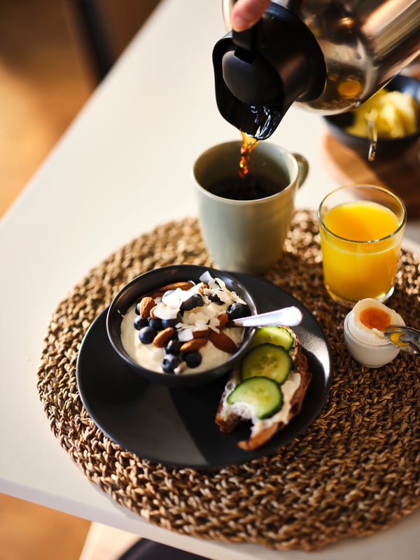 Plated breakfast food and juice with coffee being poured into a matt green FÄRGKLAR mug atop a seagrass IHÅLLIG place mat.