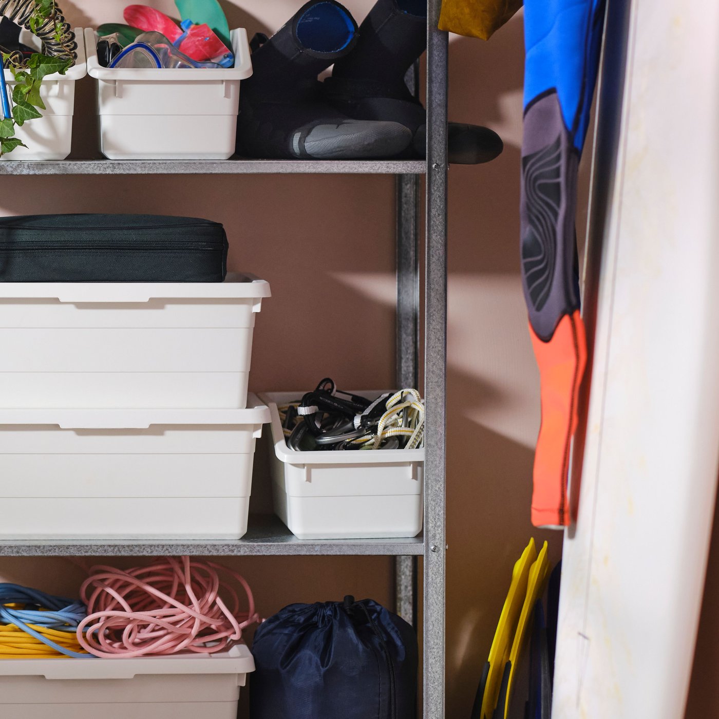 Surfing accessories and white SOCKERBIT boxes placed in a HYLLIS shelving unit next to a surfboard leaning against a wall.