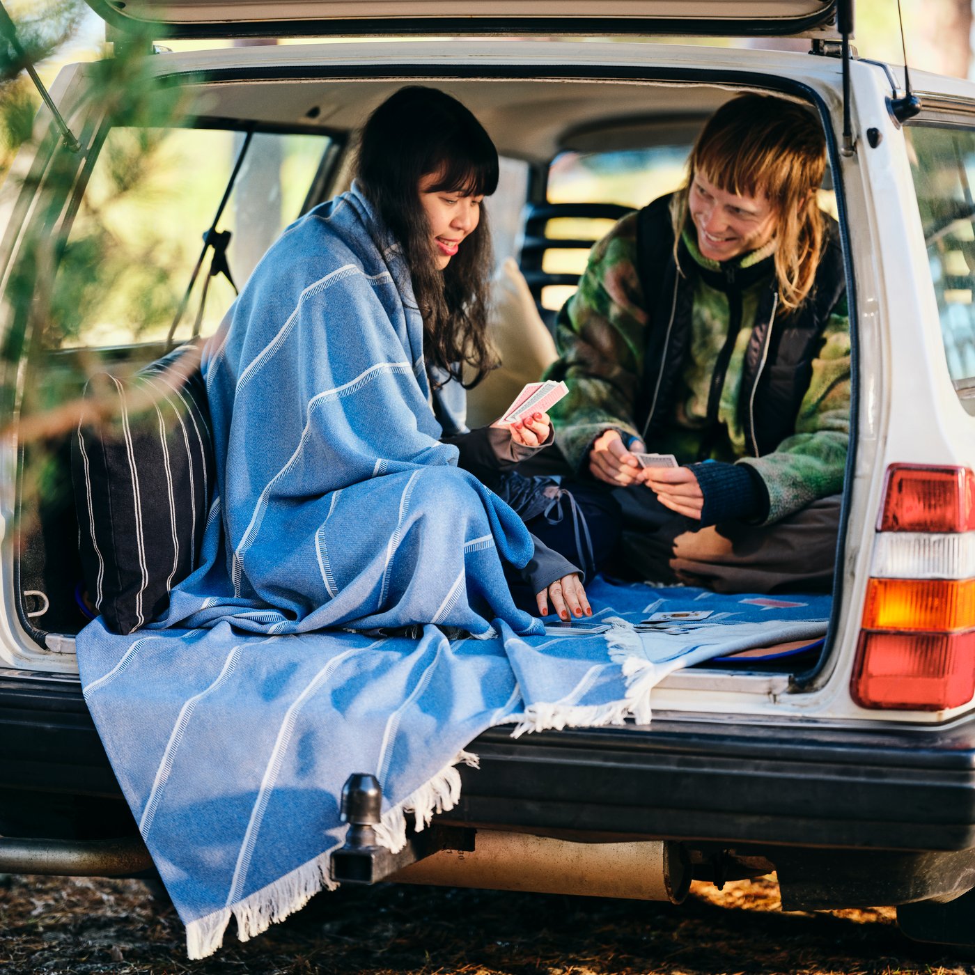 A couple is smiling and playing cards in the back of a car; one of them has a SOLUPPGÅNG throw wrapped around her shoulders.