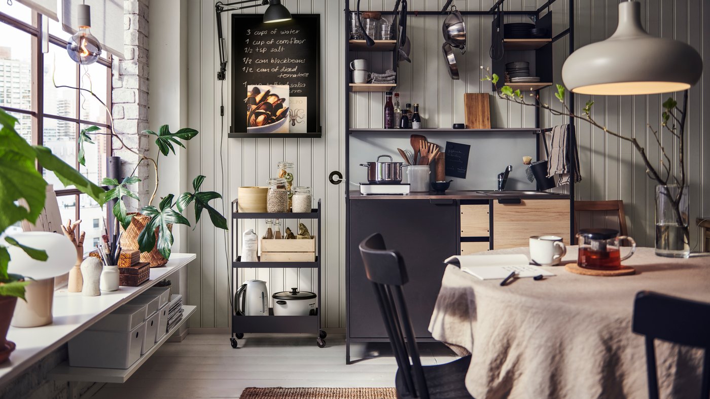 Kitchenette et table à manger dans un espace industriel, avec meubles en métal noir et bois et étagères flottantes blanches en dessous de grandes fenêtres
