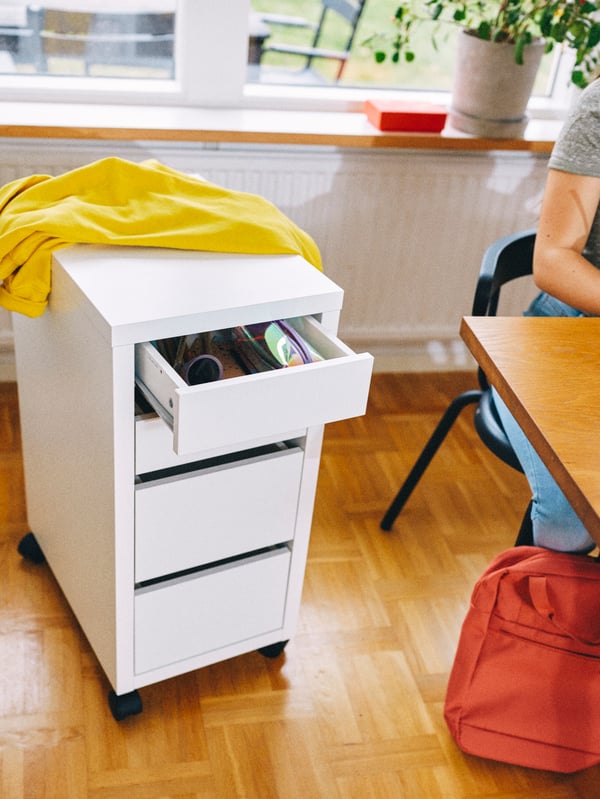 A white drawer unit on castors stands in a room with a desk. A yellow jacket is slung on top of the drawer unit.