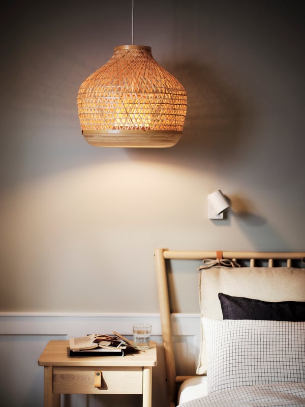 Ein minimalistisches Schlafzimmer mit einem warmen, natürlichen Touch. Eine elegante Rattan-Hängelampe sorgt für sanftes Licht, während eine dezente Wandlampe das Ambiente ergänzt. Das Bett ist schlicht, mit einem Kissen in neutralen Tönen, und ein Holztisch steht daneben, auf dem ein Glas Wasser und ein geöffnetes Buch liegen