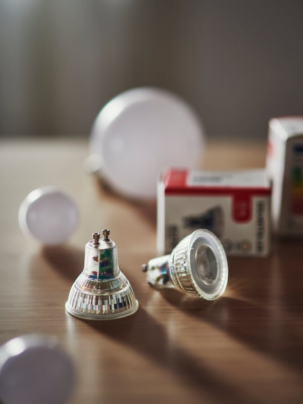Two SOLHETTA LED bulbs on a brown wooden table. Out of focus in the background are the bulb packages and some large bulbs.