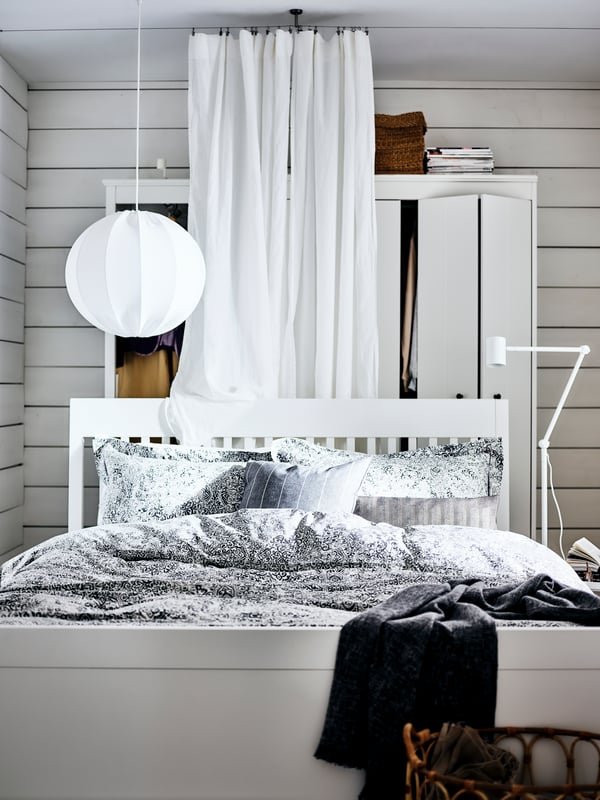 A white IDANÄS bed in a white bedroom with a curtain hanging behind, a pendant lamp above it, covered with grey bed linen.