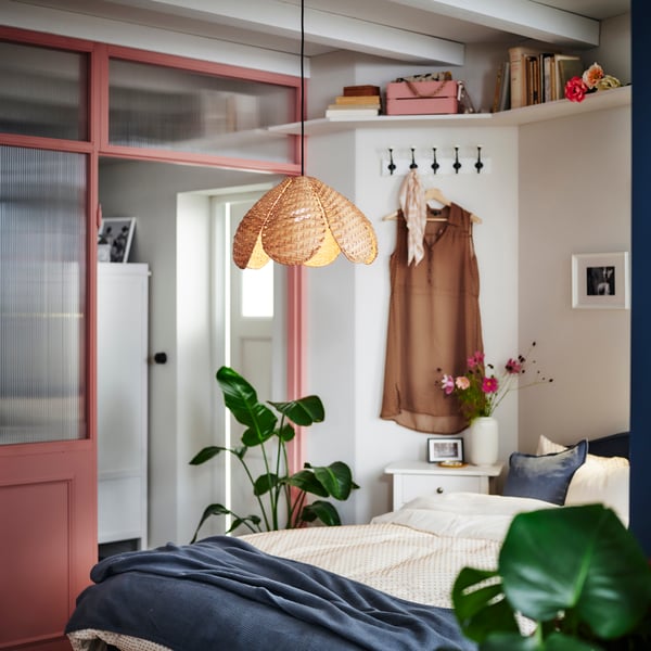 A light bedroom with leafy plants, colourful accents, clothing on a hanger and a central, lit SANDKORN rattan pendant.