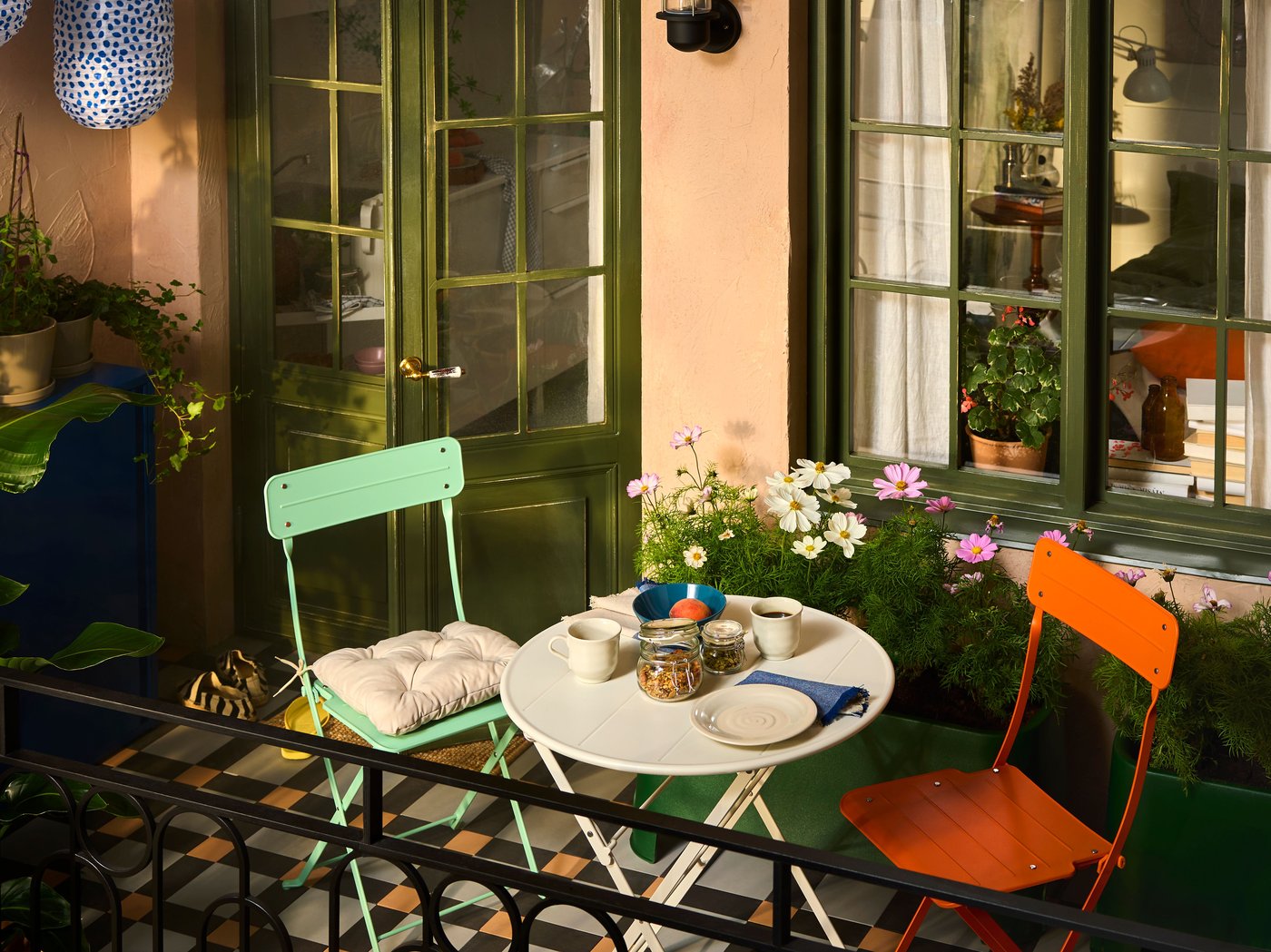 A city balcony with SUNDSÖ outdoor furniture in playful colours surrounded by plants, flowers and cabinets for storage.