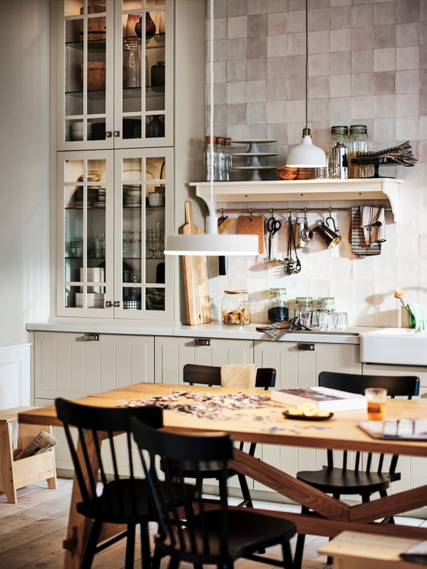 Une cuisine avec des façades de tiroir et des portes vitrées STENSUND beiges et une table MÖCKELBY en chêne autour de laquelle sont disposées des chaises NORRARYD noires.