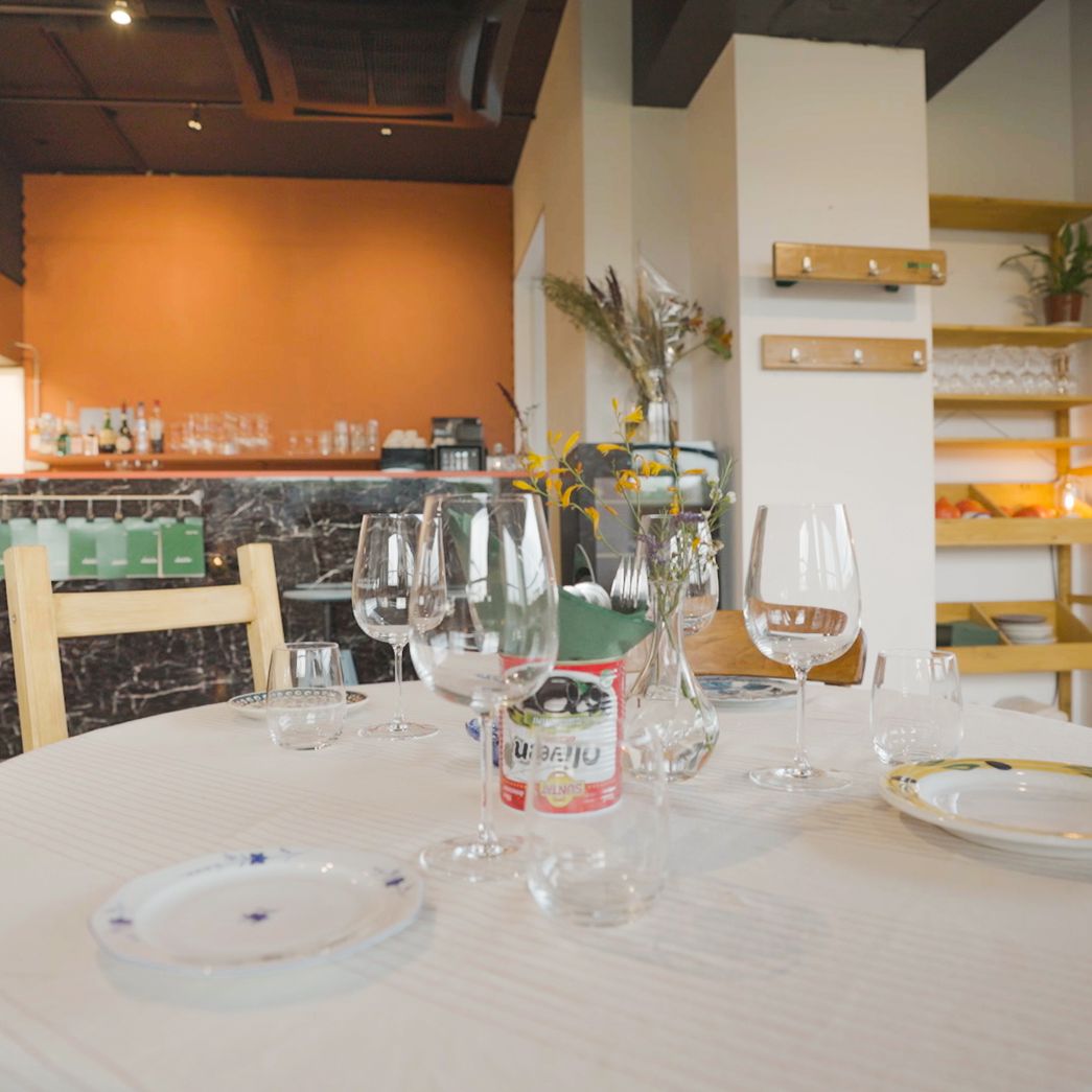Ronde tafel met glazen, borden, bloemen en tomatenblik in gezellig restaurant. Achteraan marmeren bar en warme tinten in stijlvol interieur.