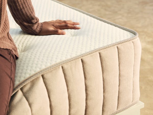 A person sitting near the corner of a medium firm/natural VATNESTRÖM pocket sprung mattress with their hand on the surface.