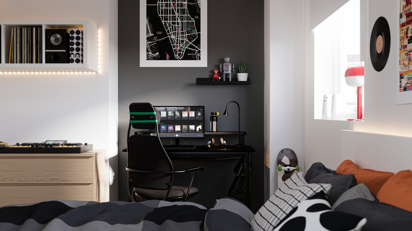 Es ist ein Schlafzimmer mit einem Bett im Vordergrund und einem Schreibtisch vor einer schwarzen Wand im Hintergrund zu sehen. Auf dem Schreibtisch steht ein Computer und davor ein Gamingstuhl.