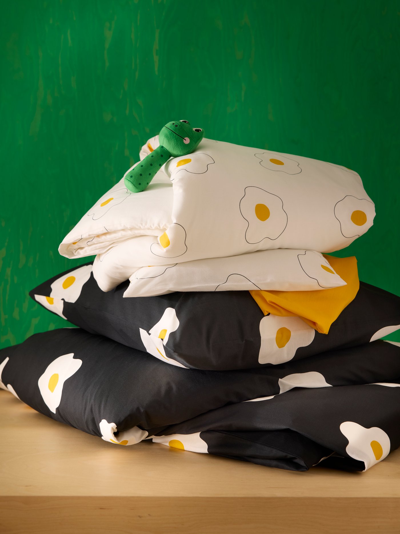 Green wall and pile of bedding including black adult bedding and white children’s bedding both with fried-egg theme.