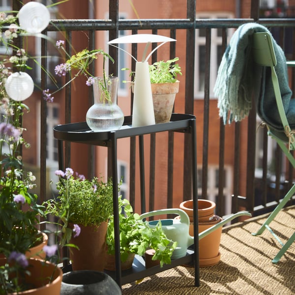 Ein kleiner Balkon in ruhiger Umgebung mit Grünpflanzen, dem Pflanzenständer OLIVBLAD in Schwarz und der Lampe SOLVINDEN.