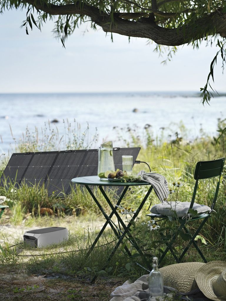 Dans un cadre extérieur paisible, un ensemble de jardin IKEA est installé à côté d'un panneau solaire, près de l'eau, à l'ombre d'un arbre.