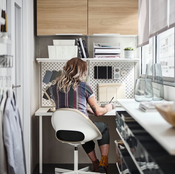 A person sitting in a beige/white swivel chair by a white BOAXEL/LAGKAPTEN shelving unit with table top on a balcony.