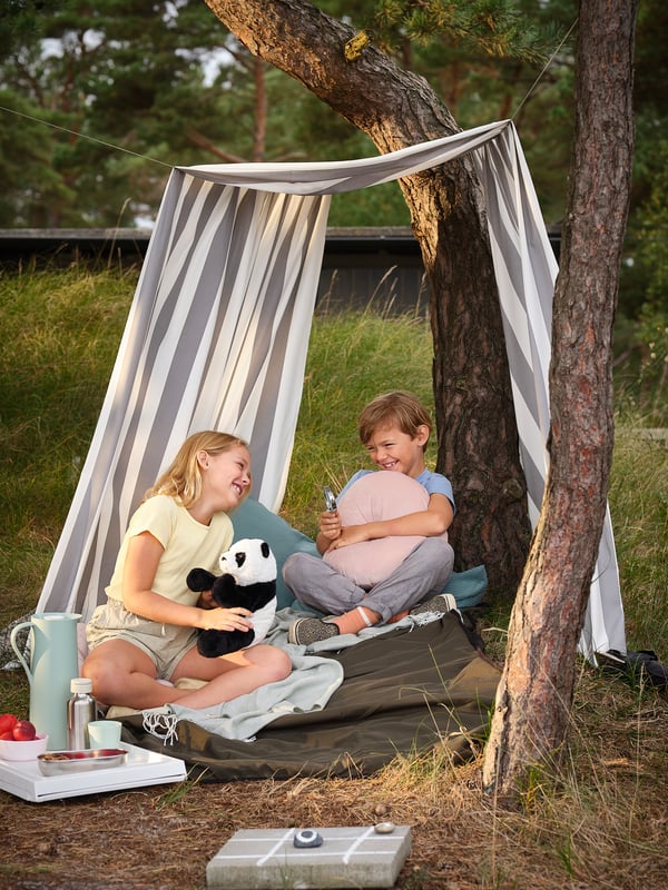 A boy hugging a KRANSBORRE cushion and a girl with a KRAMIG soft toy laugh together as they sit on a FJÄLLMOTT picnic blanket.