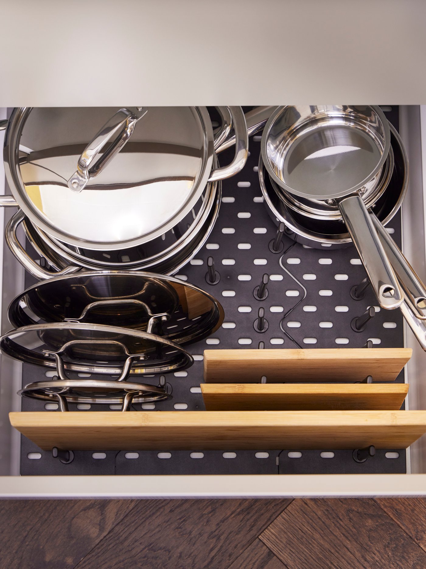 Un tiroir de cuisine bien rangé contient plusieurs casseroles et poêles, dont une grande marmite avec couvercle, une petite casserole, plusieurs poêles empilées et deux planches à découper en bois sur un fond perforé.