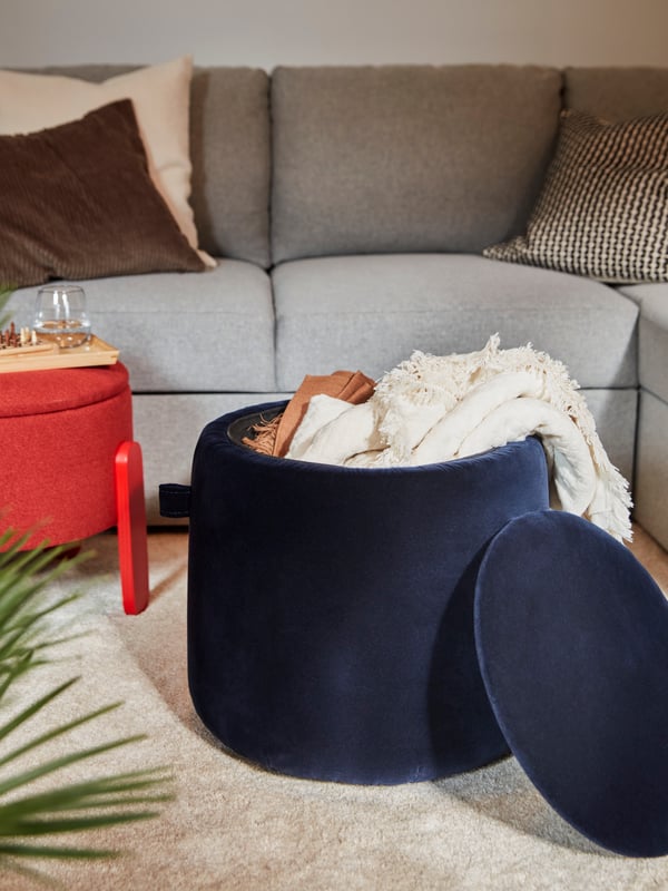 A red FÖRLUNDA pouffe with storage and a dark blue JÄRRESTAD pouffe with storage containing a throw stand near a sofa.