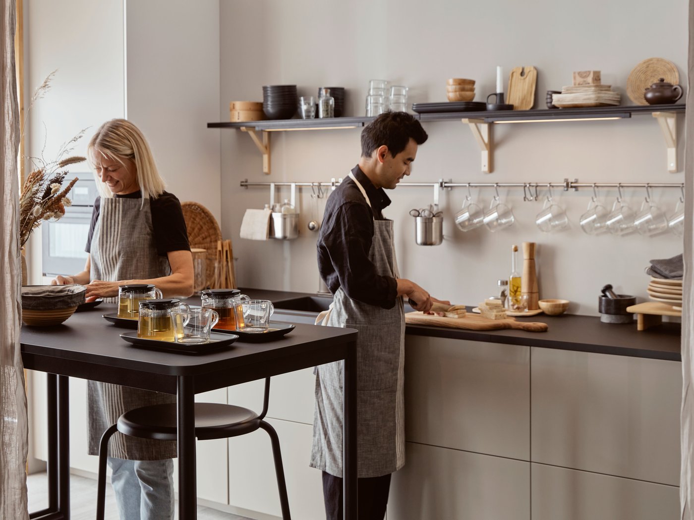 Homme debout à un comptoir de cuisine, coupant des aliments sur une planche à découper PROPPMÄTT tandis qu’une femme prépare des assiettes à une table haute avec des théières en verre au premier plan.