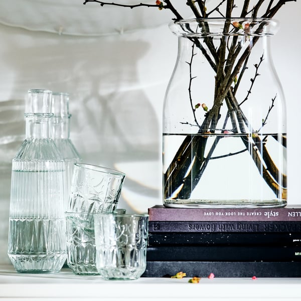 A BEGÄRLIG vase filled with water and branches on a shelf with other glasses and vases, in front of a wall with plates on it.