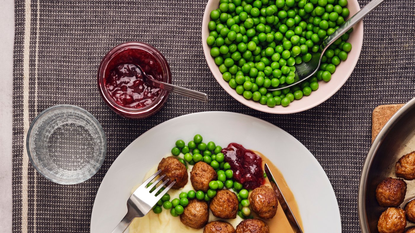 En portion kødboller, kartoffelmos og grønne ærter på en hvid tallerken med en krukke med tyttebær og et glas ved siden af.