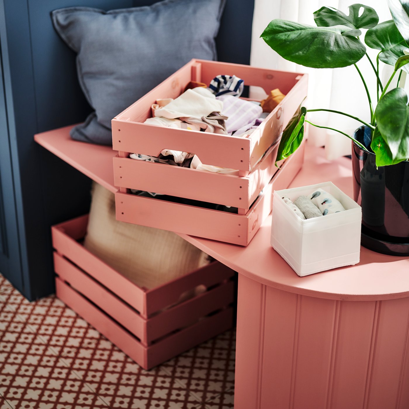 A pink-painted KNAGGLIG box on top of a bench filled with crumpled clothes beside a SKUBB box with folded clothes inside.
