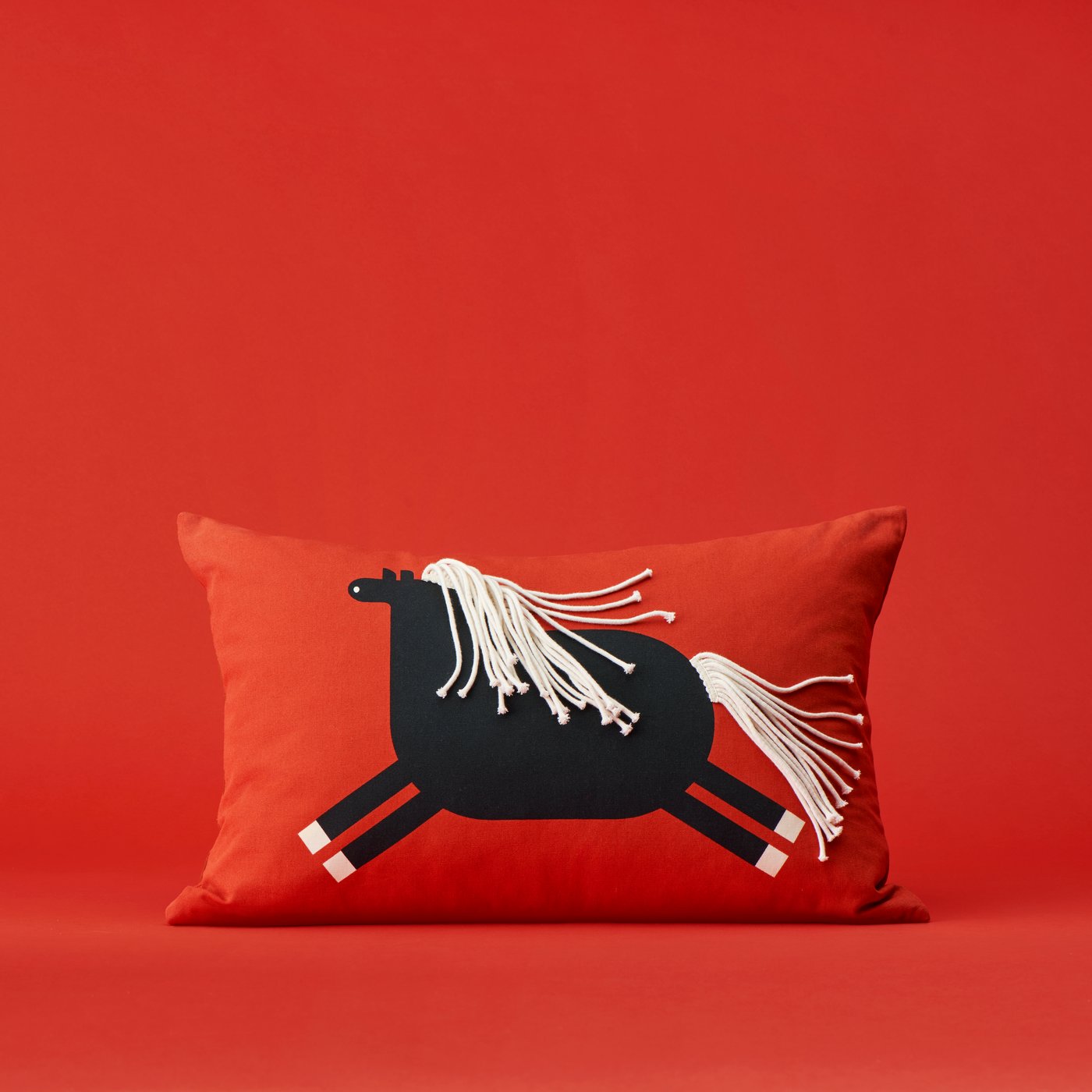 A decorative red FÖSSTA cushion cover featuring a black horse design with white tassels, against a red background.