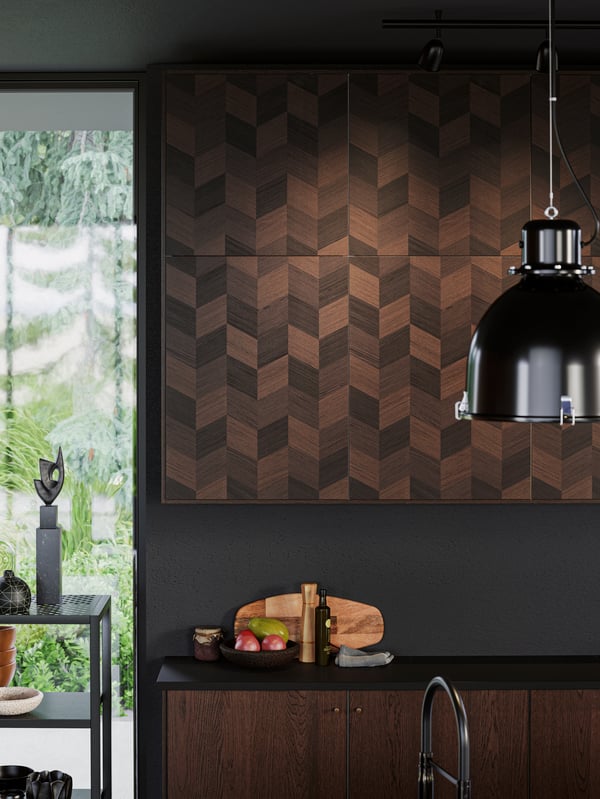 Houten kasten aan de muur met een visgraatmotief op de deuren, een zwarte hanglamp, een fruitschaal op het zwarte werkblad.