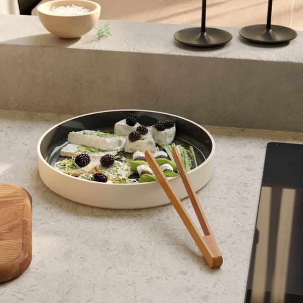 Une assiette de service OMBONAD gris foncé contient une salade de fromage avec des baies et une pince de service OSTBIT en bambou.