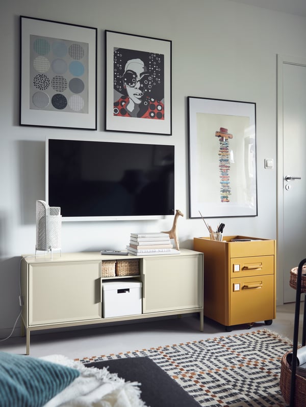 A TV and pictures are on the wall above a beige TULLSTORP metal cabinet with open storage holding baskets and a box.