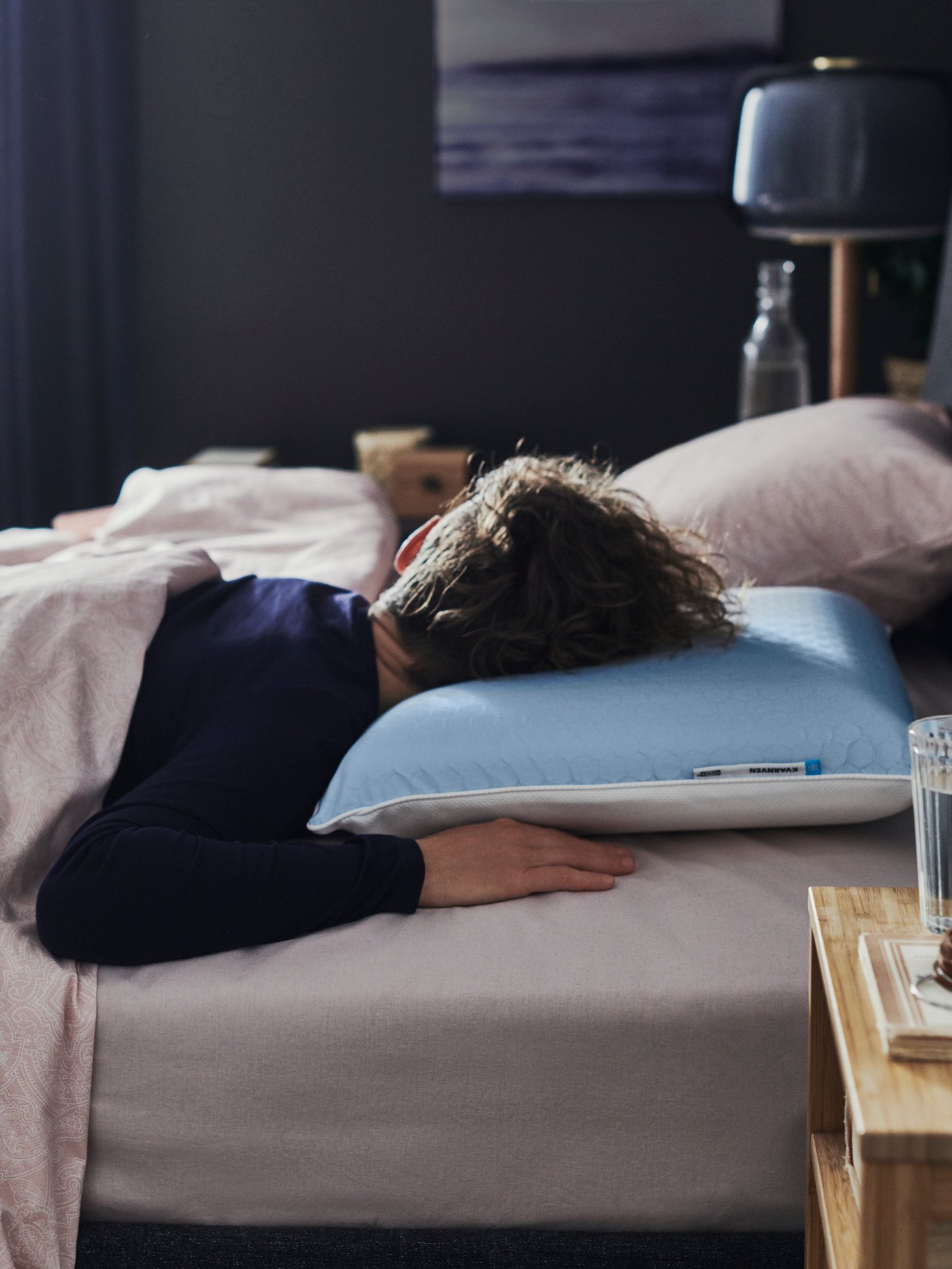 A person sleeps on a bed with a KVARNVEN ergonomic pillow designed for stomach sleepers, in a softly lit bedroom.