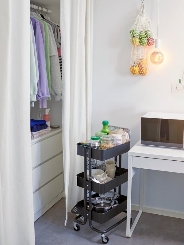 Carrello RÅSKOG nell'angolo di una stanza universitaria con pentole, piatti e alimenti in contenitori. Sulla parete è appesa della frutta - IKEA.