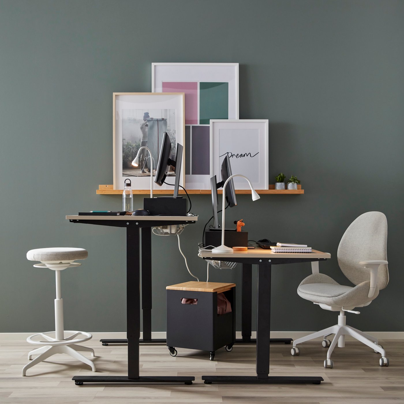 Two IKEA TROTTEN sit-stand desks in oak and black, a beige desk chair and an oak shelf on a green wall.