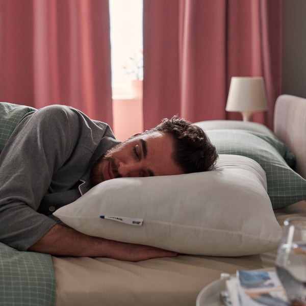 Un hombre recostado en la cama con su cabeza sobre una almohada LAPPTÅTEL. Detrás se ve una ventana con las cortinas cerradas.
