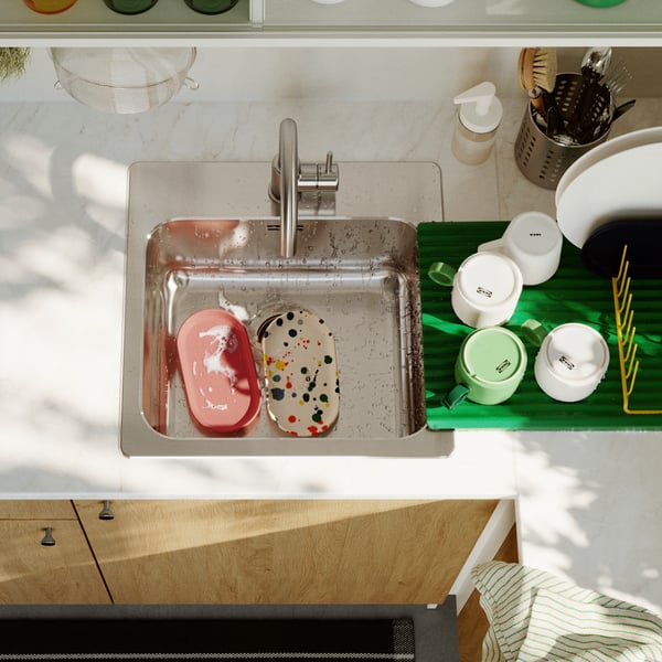A clean kitchen featuring a stainless steel LÅNGUDDEN inset sink with a colourful dish set drying beside fresh fruits.