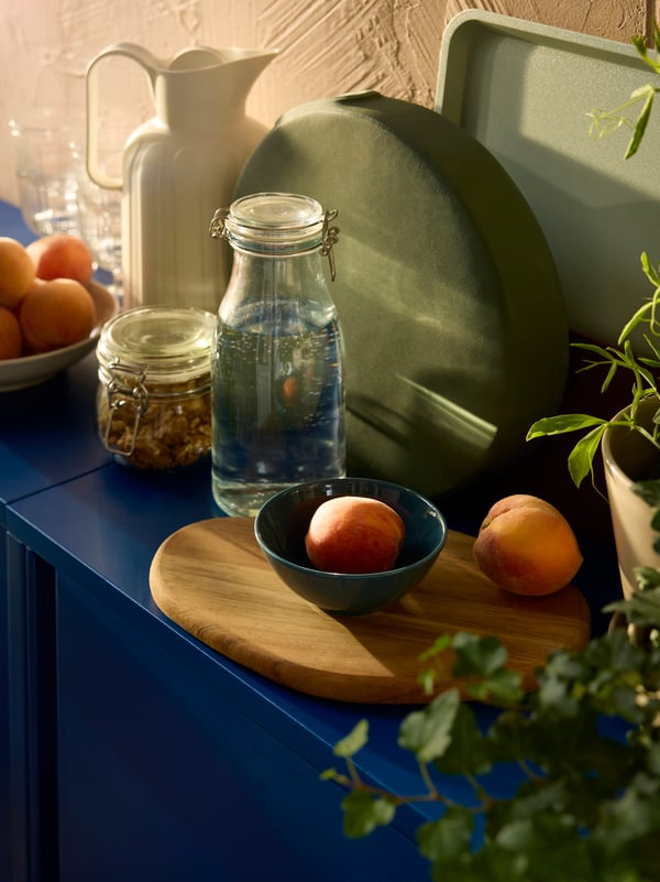 Deux bocaux KORKEN, un pichet isotherme TAGGÖGA en blanc cassé, des pêches et une planche à découper sont disposés sur une armoire SUNDSÖ bleu foncé.
