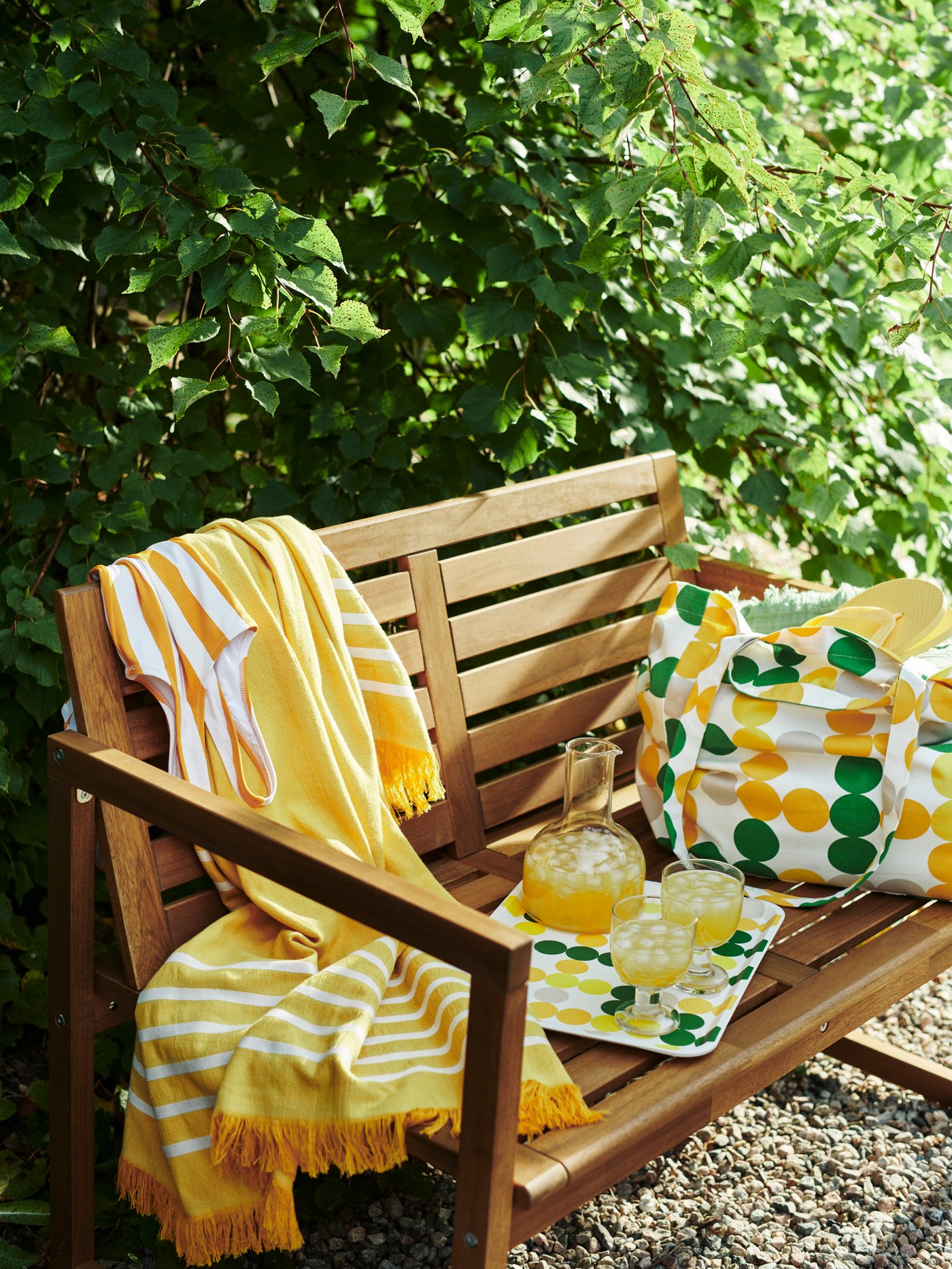 A yellow BRÖGGAN bath sheet and a dot-patterned BRÖGGAN beach bag, tray and carafe on an outdoor bench next to a lush hedge.