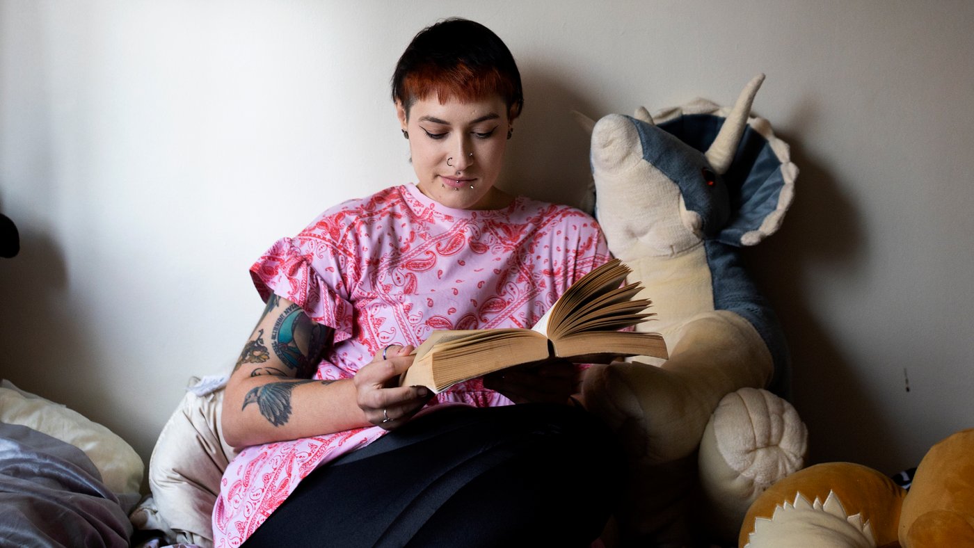 Woman sitting on her bed, surrounded by soft toys, reading a big book.